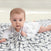 A baby lies on their stomach on an aden + anais boutique silky soft swaddle blanket with a patterned design. Wearing a white onesie adorned with light stars, the baby gazes forward curiously, gripping a corner of the swaddle. The softly blurred background adds warmth to this serene scene.