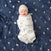 A happy baby is wrapped in an aden + anais boutique silky soft swaddle blanket featuring animal drawings, wearing a gray hat, and lying on a dark blue moon-patterned blanket.