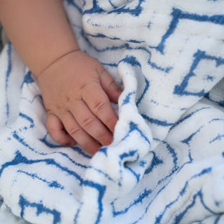 A babys hand gently grasps an aden + anais boutique silky soft swaddle blanket featuring a blue geometric pattern, highlighting the small, delicate fingers and bamboo-infused viscose fabric.