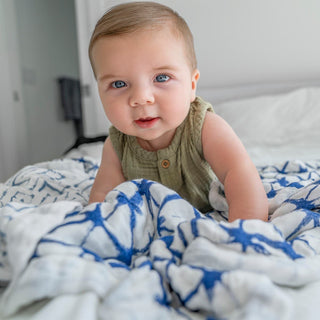 A baby with light brown hair and blue eyes is crawling on a bed, wearing a green sleeveless top. The soft, bamboo-infused viscose blanket beneath, featuring a blue abstract pattern, is from the aden + anais boutique silky soft swaddle blanket 3 pack. The blurred background adds warmth to the scene.