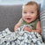 A smiling baby with short, light brown hair sits on a gray couch wrapped in HALOs Tried & True Bundle. The black and white patterned blanket complements the babys green sleeveless outfit as they look directly at the camera.