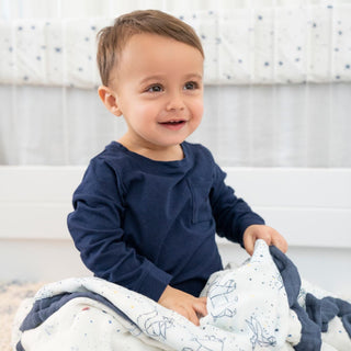 A toddler in a navy blue shirt sits on the floor, holding an aden + anais boutique silky soft muslin dream blanket with white and blue patterns. The cozy setting includes a crib with a starry design, and the toddler smiles, looking slightly to the left.