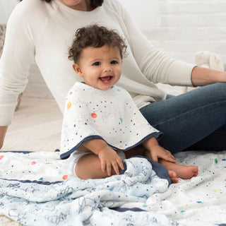 A smiling baby with a patterned bib sits on a boutique silky soft muslin dream blanket by aden + anais, featuring star and planet designs. An adult in a white shirt is nearby in the bright, cozy room, creating a cheerful and relaxed atmosphere ideal for bamboo-infused viscose comfort.