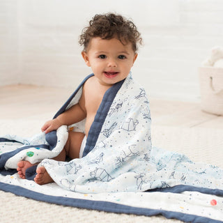 A baby with curly hair sits on a rug, wrapped in an aden + anais boutique silky soft muslin dream blanket. The cozy bundle is smiling at the camera in a soft, neutral-colored room with a plush toy in the corner.
