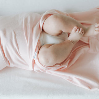 A baby lies on a soft white surface, partially covered by a HALO Swaddle 100% Cotton TOG 1.5. The adjustable wings secure them as their legs and feet are exposed, with one foot playfully touching the other leg.