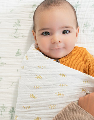 A baby with big brown eyes, wearing a mustard yellow top, is swaddled in a white blanket with yellow floral patterns and lying on a patterned sheet. An adults hand gently holds the baby.