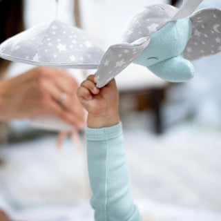A child in a light blue sleeve reaches for a soft toy elephant with star-patterned fabric hanging from an aden + anais baby activity gym, while a blurred adult hand in the background hints at sensory development play.