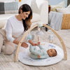 A woman smiles at a baby on an aden + anais baby activity gym, which includes a circular play mat and birchwood frame with soft hanging toys for sensory development. The cozy room features woven baskets and cushions in the background.
