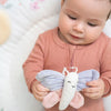 A baby, dressed in a pink outfit, lies on a soft, light-colored surface while holding a stuffed butterfly toy from aden + anaiss eco-friendly baby activity gym. The toy features pastel colors with purple and pink wings and a white body.
