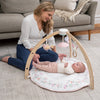 A woman sits on the living room floor, smiling at a baby on an aden + anais baby activity gym. The wooden play arch above features eco-friendly materials and hanging toys, including a pink bird and rabbit, with a gray couch in the background.