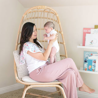 A woman in a white shirt and pink pants sits on a wicker chair, cradling her baby in a white onesie. On her shoulder is an aden + anais boutique 100% cotton muslin burpy bib, highlighting its practicality. Behind them, shelves filled with colorful books add vibrancy to the cozy setting.
