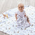 A baby in a white onesie sits on a patterned animal blanket, holding an aden + anais boutique cotton muslin dream blanket. A giraffe toy lies nearby on the wooden floor.