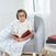 A toddler with round glasses sits barefoot in a gray armchair, holding an open book resembling Harry Potter. Dressed in white, the child is perched on a boutique iconic cotton muslin dream blanket by aden + anais. Soft light filters through sheer curtains behind them.