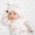 A baby in a white snuggle knit™ newborn gift set with pink floral patterns and a matching knotted hat from aden + anais rests on a white surface with pink dots, gazing upwards. The left hand near the head perfectly captures the charm of this adorable ensemble.