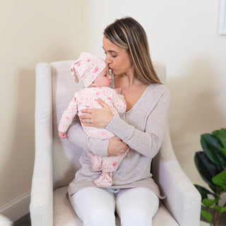 A woman in a light gray sweater and white pants kisses a baby dressed in aden + anais comfort knit™ newborn gown + hat gift set, featuring matching pink floral pajamas. She sits in a beige armchair with a green plant visible, creating a calm, cozy setting.