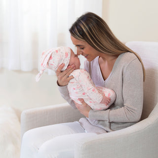 A woman with long hair, in a light gray sweater, sits on a chair holding a baby in an aden + anais comfort knit™ newborn gown + hat gift set. They touch noses and smile in a softly lit room with a white curtain.