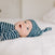 A baby in a snuggle knit™ newborn gift set by aden + anais lies on a white bed, its blue and white stripes and matching knotted hat creating a serene scene. The babys content gaze and open mouth add charm, while the softly blurred background captures the precious moment beautifully.