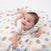 A smiling baby lies on a colorful Disney™ boutique swaddle blanket with circular designs in blue, brown, and yellow. The baby is wearing a white onesie and gazing upward while wrapped in a soft aden + anais 100% cotton muslin swaddle blanket.