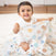 A baby in a white onesie sits on a white rug, smiling with a Disney™ boutique by aden + anais cotton muslin swaddle blanket featuring colorful circular designs. The wooden floor and blurred furniture create an ideal backdrop for playtime with their favorite swaddle blanket.