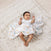 A baby joyfully raises its arms while lying on a textured blanket, wearing a white onesie and partially covered by an aden + anais Disney™ boutique 100% cotton muslin swaddle from the 3 pack, surrounded by colorful patterns.