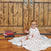 A baby, resembling Harry Potter with round glasses, sits on a wooden floor by a rustic door wrapped in an aden + anais boutique iconic 100% cotton muslin swaddle adorned with red and blue stars, with a stack of books nearby completing the charming scene.