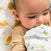 A baby lies on a bed wrapped in an aden + anais organic cotton muslin swaddle blanket featuring green and blue designs. The child, in a beige long-sleeve top, clutches the 100% organic muslin swaddle while resting on lemon-patterned bedding.