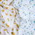 On a white background, crumpled fabric displays two prints: yellow oranges with green leaves and blue berries with green leaves. These vibrant patterns adorn aden + anais 100% organic cotton muslin swaddle blanket 2 pack, which is GOTS certified for quality and sustainability.