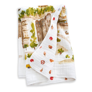 The aden + anais organic cotton muslin dream blanket features a soft, white fabric with a colorful cityscape on one side and playful prints of apples, pretzels, pizza, and ice cream on the other—perfect for babies.