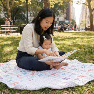 A woman reads a picture book to a toddler with a white bow while sitting on an aden + anais organic cotton muslin dream blanket in the park. Sunlight filters through the trees as people stroll by in the background.