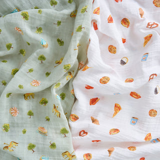 This aden + anais organic cotton muslin swaddle blanket 2 pack features one pale green carnival print and one white with colorful food prints—playful, breathable, and perfect for your baby.