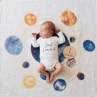 A baby in a "Just Landed" onesie lies on a planet-themed blanket, cuddled up in an aden + anais organic cotton muslin swaddle blanket (2 pack), next to a wooden "1 month" sign.