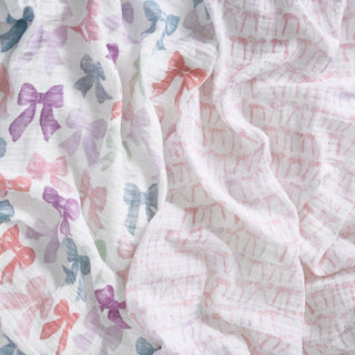 Close-up of aden + anais organic cotton muslin swaddle blanket 2 pack: one with colorful bows in pink, purple, blue, and green, and one with small light pink bows. Both feature a soft, airy texture ideal for gentle snuggles.