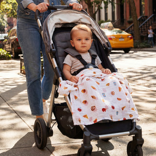 A baby sits in a stroller covered with the aden + anais organic cotton muslin dream blanket decorated with colorful food prints, while an adult pushes the stroller down a sunny, tree-lined city sidewalk.