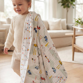 A smiling toddler holds the aden + anais essentials cotton muslin dream blanket with colorful car patterns, standing in a bright, cozy living room with neutral furniture and wooden floors.
