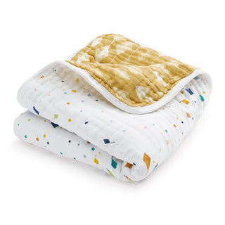 The aden + anais essentials cotton muslin dream blanket features a soft, folded design with one white side adorned with colorful geometric shapes and a mustard yellow reverse with a subtle wavy pattern.