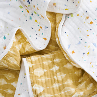 A close-up of the aden + anais essentials cotton muslin dream blanket, featuring quilted fabric with a white section adorned with multicolored confetti dots and a yellow area showcasing a subtle geometric diamond pattern.