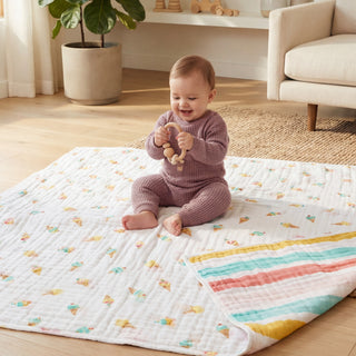 A baby in a mauve knit outfit sits on an aden + anais essentials cotton muslin dream blanket, holding a wooden toy and smiling in a bright, cozy living room with wood floors and modern furniture.