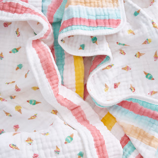 The aden + anais essentials cotton muslin dream blanket features pastel pink, yellow, and blue stripes with tiny colorful ice cream cone patterns and is shown gently crumpled.