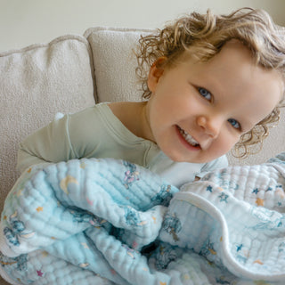 A young child with curly hair and blue eyes smiles while holding the aden + anais essentials cotton muslin dream blanket, sitting on a beige couch.