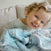 A young child with curly hair and blue eyes smiles while holding the aden + anais essentials cotton muslin dream blanket, sitting on a beige couch.