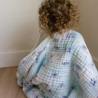 A young child with curly hair sits on a light wooden floor, wrapped in the aden + anais essentials cotton muslin dream blanket with cartoon animal and star patterns, in a simple room with light-colored walls.