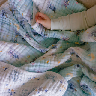 A child’s hand rests on the essentials cotton muslin dream blanket by aden + anais, featuring soft pastel colors with cartoon animals and stars. The cozy scene highlights the blanket’s gentle cotton muslin fabric, perfect for comfort and peace.