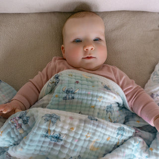 A baby with blue eyes and rosy cheeks lies on a soft surface, cozily wrapped in the aden + anais essentials cotton muslin dream blanket, which features animal and star patterns, and wears a light pink outfit.