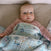 A baby with blue eyes and rosy cheeks lies on a soft surface, cozily wrapped in the aden + anais essentials cotton muslin dream blanket, which features animal and star patterns, and wears a light pink outfit.