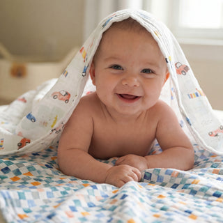 A smiling baby lies on their stomach under the aden + anais essentials cotton muslin swaddle blanket, featuring car patterns. Sunlight from a window behind creates a cozy, cheerful atmosphere. Available in a 2 pack.