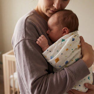 A woman gently cradles her sleeping baby, wrapped in an aden + anais essentials cotton muslin swaddle blanket, against her chest in a softly lit room.