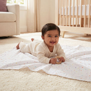 A smiling baby in a cream outfit lies on an aden + anais essentials cotton muslin swaddle blanket (2 pack)—white with colorful specks—holding a toy on a soft carpet in a cozy nursery with a crib and armchair in the background.