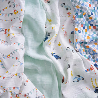 Three soft essentials cotton muslin swaddle blankets from aden + anais are shown: one with colorful pennants, one with cars and trees, and one in blue-orange checks, all displayed on a white background.