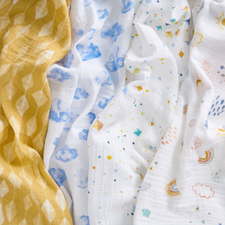 The aden + anais essentials cotton muslin swaddle blanket 4 pack includes lightweight blankets: one yellow with a leaf pattern, and three white—one with blue clouds, one with colorful shapes, rainbows, and weather icons.