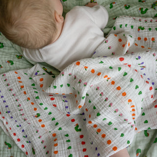 A baby in a white onesie lies on a light green patterned blanket, partially covered by the HALO The Very Hungry Caterpillar Bundle cotton swaddle decorated with dotted lines and small green, orange, and pink shapes.